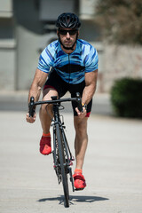 Front view of muscular young man cycling in a city looking at camera. Athletic cylcist training and riding a bike wearing helmet and sunglasses in the street.