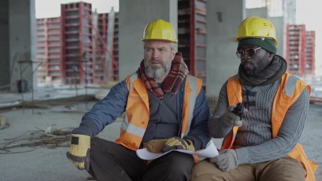 Medium shot of two ethnically diverse builders wearing safety vests and hard hats sitting on concrete floor of unfinished construction site and having discussion