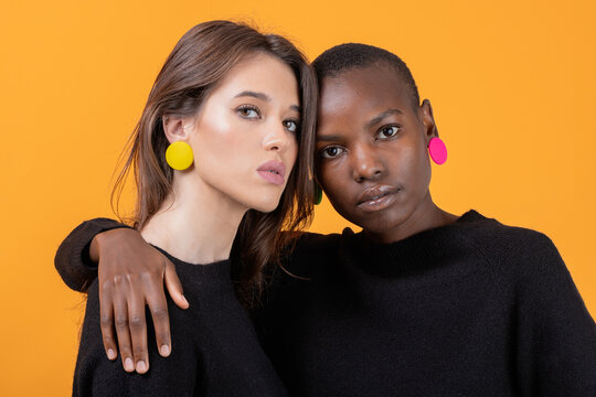 Diverse Young Female Friends Wearing Colorful Earrings Embracing Each Other Against Yellow Background