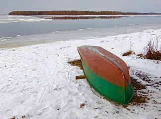 An overturned duralumin boat on a snow-covered river bank in late autumn, at the beginning of...