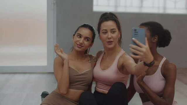 Pan shot of smiling multicultural girls taking selfie on smartphone while waiting for fitness class beginning