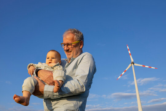 From below smiling elderly man holding a baby with a wind turbine in the background under a clear blue sky