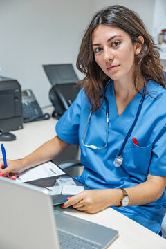 Young woman doctor sitting at table with laptop smartphone and making note on clipboard
