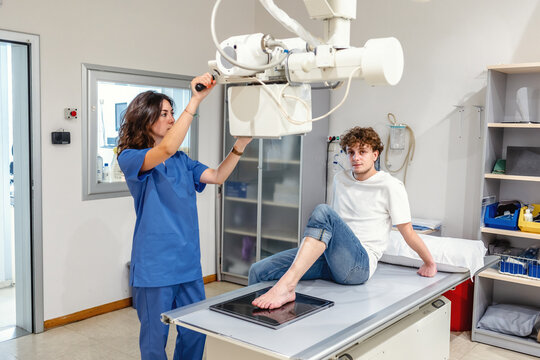 Young female technician adjusts X Ray machine for scanning patient sitting on bed in clinic