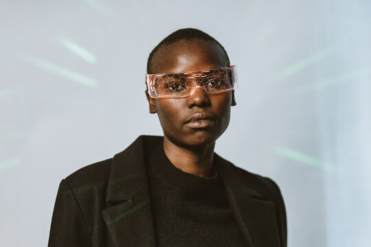 Portrait of confident young bold African American woman in smart futuristic VR glasses and long black trench coat standing looking at camera against neon light illuminated background