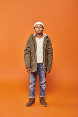 Fototapeta premium preadolescent african american boy in winter attire with beanie hat posing on orange backdrop