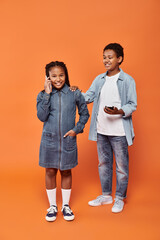 happy african american girl and boy in casual wear using smartphones on orange background