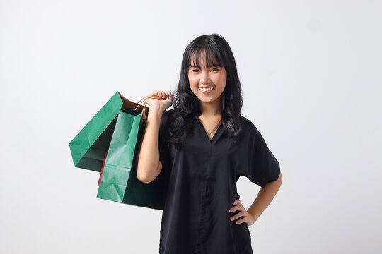 Portrait of excited Asian woman in black shirt holding and showing colorful paper shopping bags. Shopaholic girl and discount buying concept. Isolated image on white background