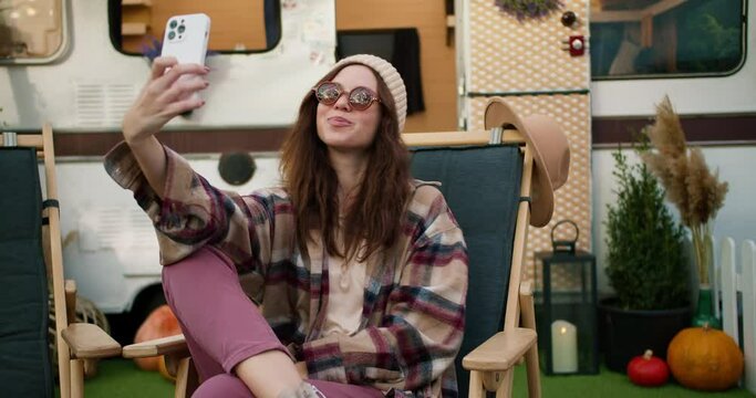 A happy girl in a white hat and plaid shirt sits on a green chair against the backdrop of a picnic trailer and takes a selfie using a white smartphone during her trip outside the city in the summer