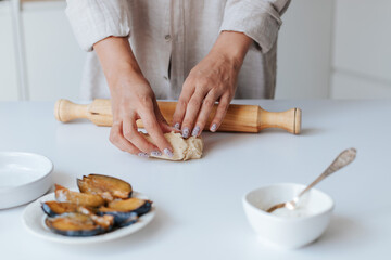Female hands knead dough on a white table in a bright kitchen. next to it is a rolling pin, a plate with chopped plums, a bowl of cream cheese and a baking dish