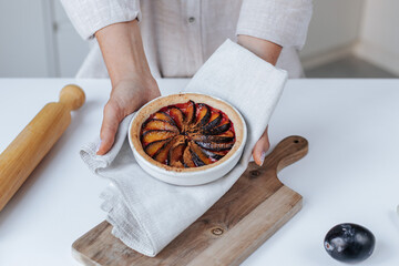 female hands hold a mold with plum pie in a light kitchen. on the table there is a rolling pin,...