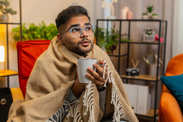 Sick Indian man in plaid sit alone shivering from cold on chair drinking hot tea in unheated...