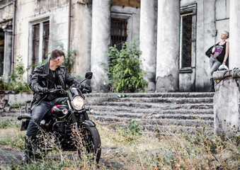 male motorcyclist in a black leather jacket and blue jeans looks at the girl