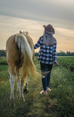 young blonde girl in a hat and a plaid shirt walks with a horse on a farm in the village