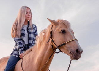 Obraz premium young blonde girl dressed in blue jeans and a plaid shirt sits on a horse at the ranch close up