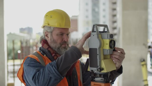 Waist up of professional middle-aged Caucasian engineer in yellow hardhat using total station while working at unfinished construction site