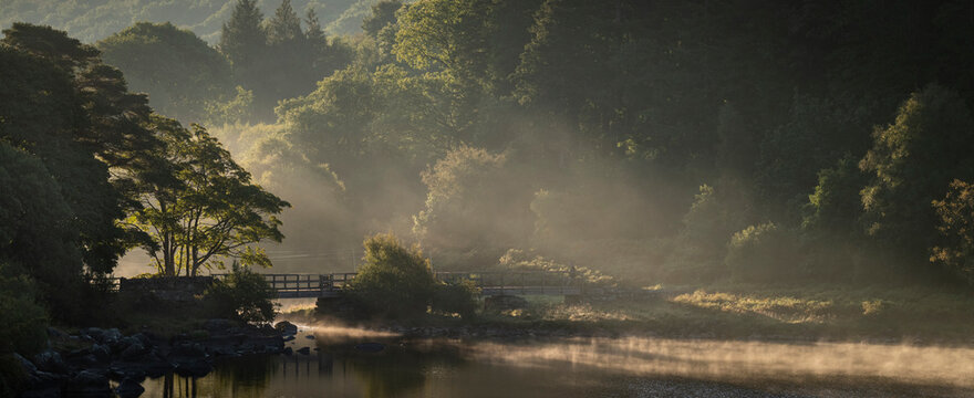 Footbridge crossing Nant Gwyd and Llynnau Mymbyr at sunrise with morning mist, Capel Curig, Snowdonia National Park, Wales