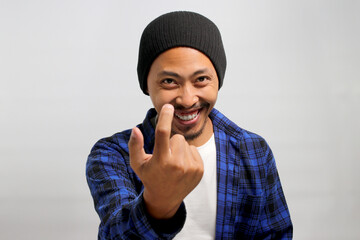 Happy, smiling young Asian man, dressed in a beanie hat and casual shirt, invites with a beckoning COME HERE gesture while standing against a white background.