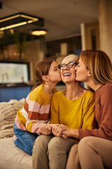 Three generations, focus on the smiling grandma, getting kisses from her grandchild and her daughter.