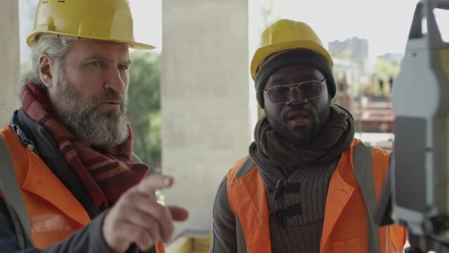 Chest up of African American workman speaking to middle-aged Caucasian male contractor, standing by total station at construction site