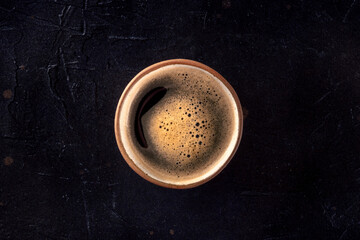 A cup of black coffee with froth, overhead flat lay shot on a dark background