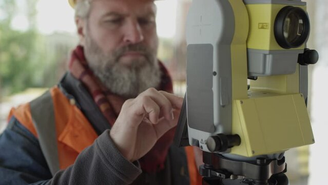 Waist up of grey haired bearded Caucasian mature engineer in hard hat adjusting total station while working at construction site