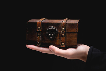 The hand of a young Caucasian woman holds a small wooden chest on a black background. Low key
