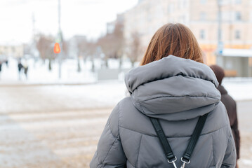 Back view of a female pedestrian in winter clothes standing by the road at a traffic light - obrazy, fototapety, plakaty