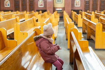 Selective focus view of cute little girl wearing pink winter clothes sitting in church pew and...