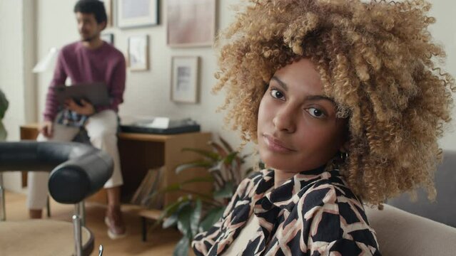 Portrait of young curly-haired black businesswoman in casualwear taking off eyeglasses and smiling on camera while posing during workday in office