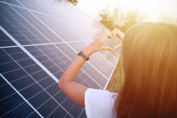 Pretty young girl staying near solar panels and showing heart symbol. Solar energy concept....