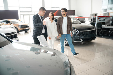 Young couple talking to sales person in car showroom choosing automobile