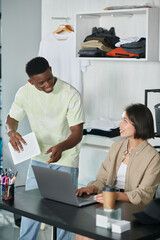 african american designer with digital tablet talking to asian woman near laptop in print studio