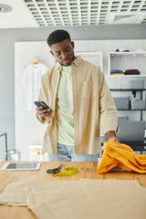 african american designer with smartphone touching fabric samples near scissors and digital tablet