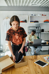 asian woman packing garments in carton box near african american designer at laptop in print atelier