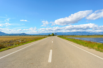 A straight road leading towards mountains under a bright blue sky with clouds, view of lonely road in the ebro delta, tarragona, catalonia, spain