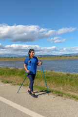 Outdoor enthusiast with walking poles beside a tranquil water body on a bright day, Hispanic Latina woman walking with trekking poles in the Ebro Delta natural park, Tarragona, Catalonia, Spain