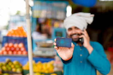 Young Indian fruit seller showing debit or credit card and phone at the camera, mixing tradition...