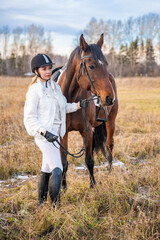 Pretty blond professional female jockey standing near brown horse in field in winter. Friendship with horse