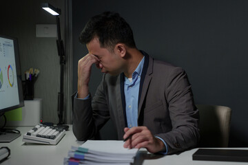 A young Asian businessman experiences stress, headaches, eye strain, and fatigue while working on a PC. Stack of financial documents on the desk feeling sick Office syndrome concept.