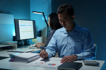 Young Asian businessman holds a pen and points to a chart graph on a document to analyze market data, finance, balance sheet, net profit, taxes to plan an online sales strategy startup business idea.