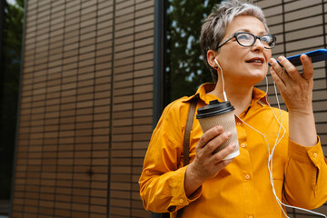 Senior woman recording voice message via smartphone and drinking coffee while standing outdoors