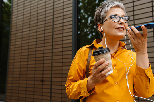 Senior woman recording voice message via smartphone and drinking coffee while standing outdoors - Powered by Adobe