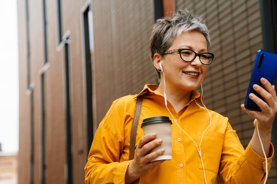 Smiling senior woman using mobile phone and drinking coffee while standing outdoors