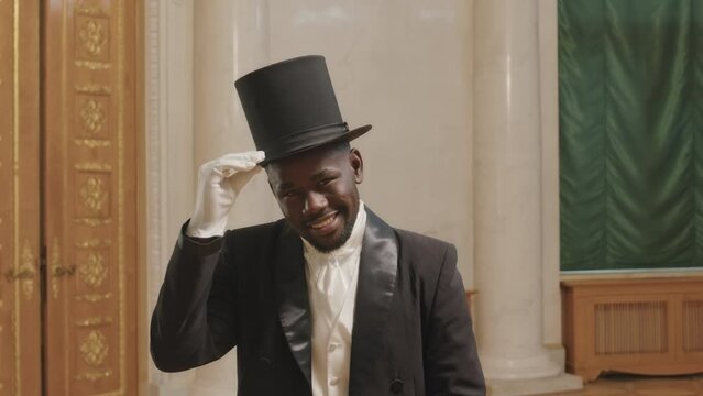 Zoom in slowmo portrait of smiling young Black man wearing tuxedo, black cylinder hat and white gloves posing for camera in classical style ballroom with wooden parquet floor and doors
