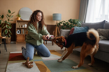 Full length portrait of happy young woman playing with dog sitting on floor in cozy home interior,...