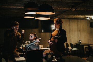 Pregnant employee gently touching her stomach while working and discussing with her teammates at modern office setting during late night work shift.
