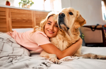 Love to animal. Happy blonde woman cuddling furry pet while lying on soft bed under checkered blanket. Pedigree golden retriever and female owner enjoying home comfort together in strong embrace.