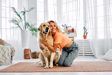 Heartwarming connection. Joyful lady in casual wear snuggling her cherished purebred dog on floor at bright living room. Caucasian woman with blond hair spending leisure with canine buddy.