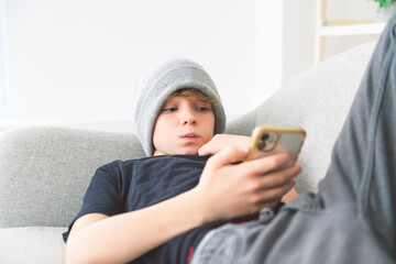 young boy lay on the couch at home watching cellphone wearing toque - obrazy, fototapety, plakaty